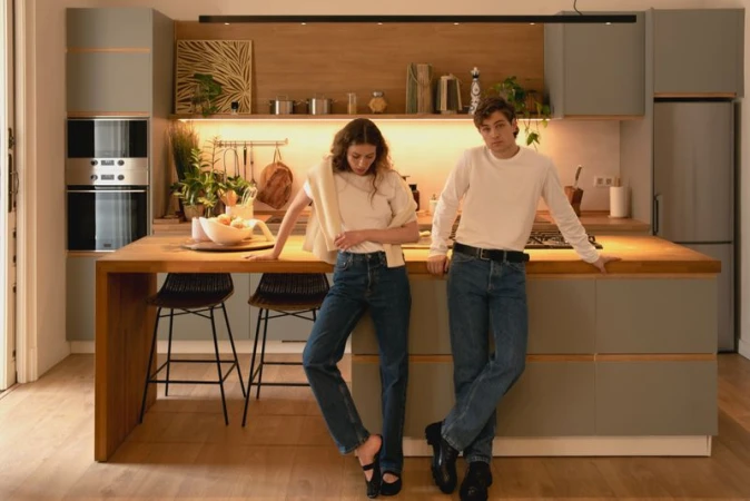 Young couple poses casually in a modern kitchen with gray cabinets, a butcher block island, bar stools, and stainless steel appliances.