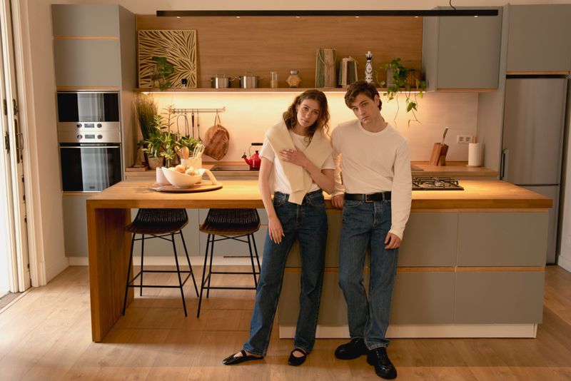 Young couple in casual clothes poses in a modern kitchen with light gray cabinets and wooden countertops, illuminated by natural light.