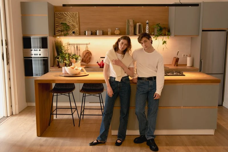 Young couple in casual clothes poses in a modern kitchen with light gray cabinets and wooden countertops, illuminated by natural light.