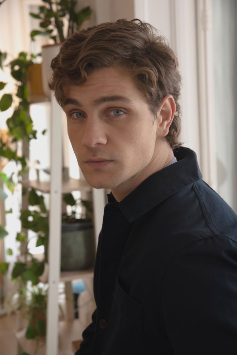 Portrait of a handsome adult male with curly brown hair and blue eyes, wearing a collared shirt, indoors with plants in background.