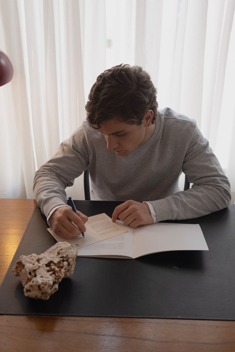 Adult male seated at desk, writing on paper with pen, indoors with natural light. Rock and curtain visible.
