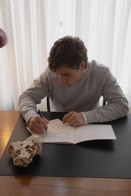 Adult male seated at desk, writing on paper with pen, indoors with natural light. Rock and curtain visible.
