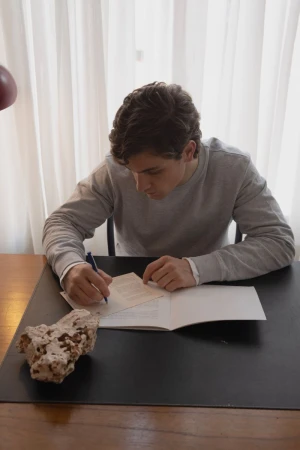 Adult male seated at desk, writing on paper with pen, indoors with natural light. Rock and curtain visible.