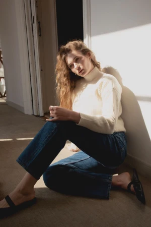 Woman with long curly hair wearing a cream turtleneck sweater and blue jeans sitting on the floor leaning against a white wall.