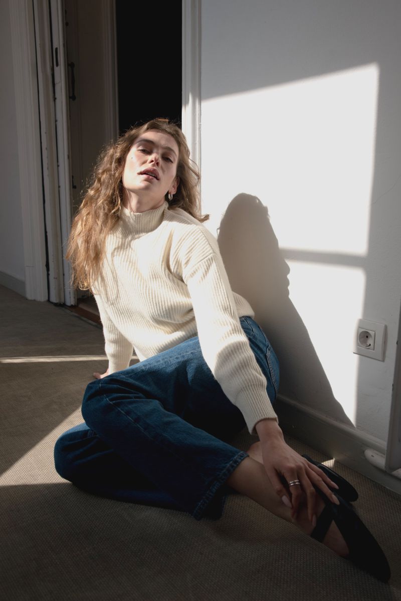A young adult female model with long wavy hair sits on the floor in a brightly lit room wearing casual jeans and a cream sweater.