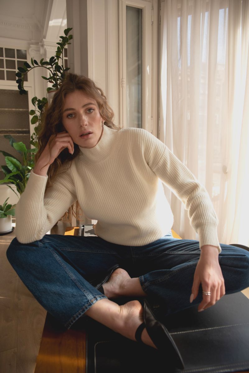 A young woman in a cream-colored sweater and blue jeans sits cross-legged indoors, looking at the camera with a neutral expression.