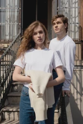 A man and a woman in white t-shirts standing outdoors on stone stairs holding a cream knit sweater