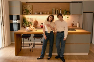 A young man and woman wearing white t-shirts and blue jeans posing in a modern kitchen with wooden countertops.