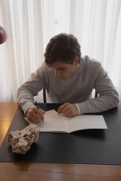Young man in a grey crewneck sweatshirt seated at a dark desk writing in an open notebook near a raw stone piece.