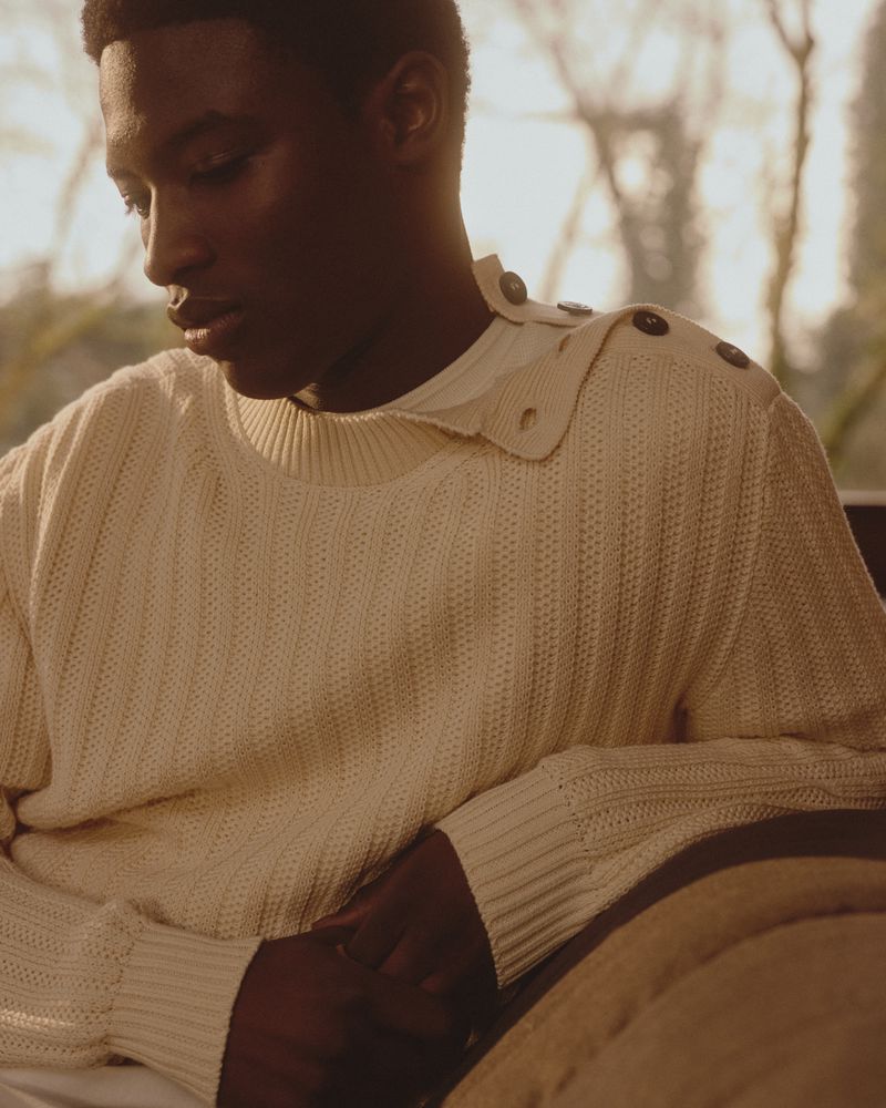 Portrait of a young African male model wearing a cream-colored knit sweater with button details, softly lit against a natural outdoor background.