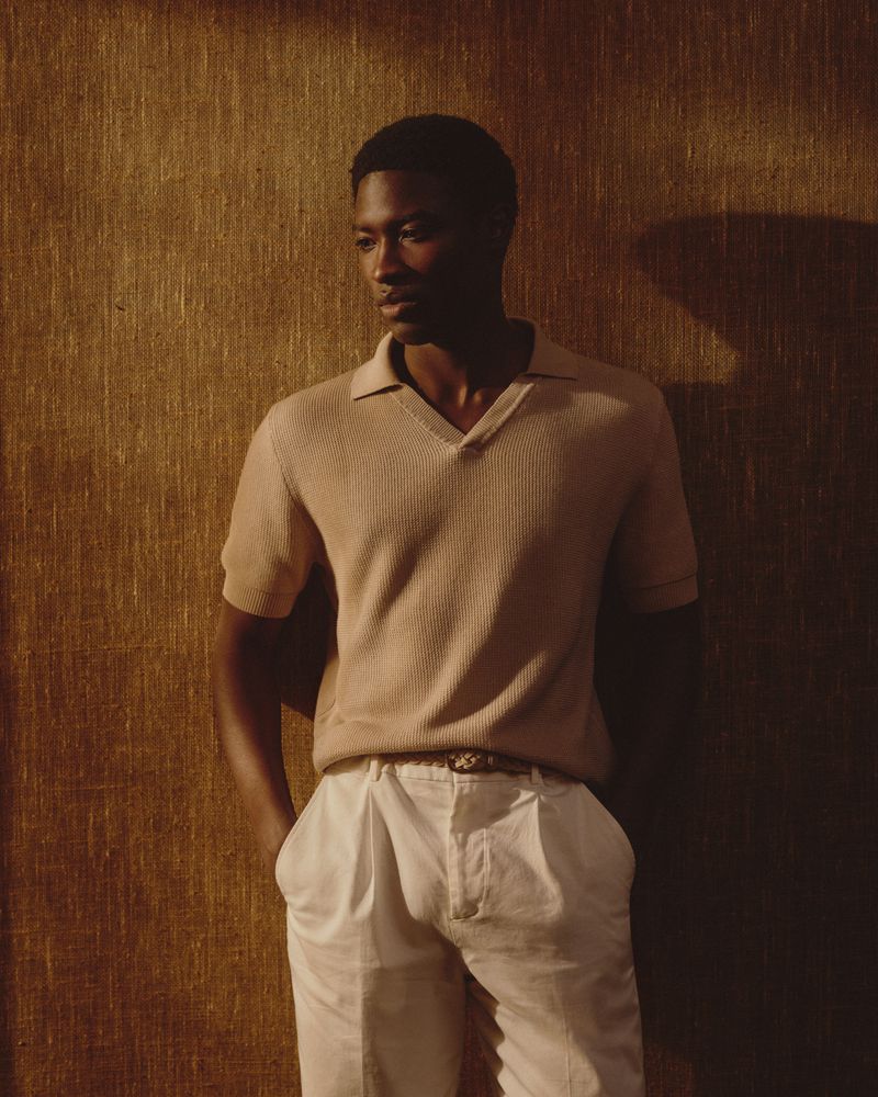 Stylish adult Black man in a beige knit shirt and white slacks posing in front of a neutral wall in a fashion editorial shot.