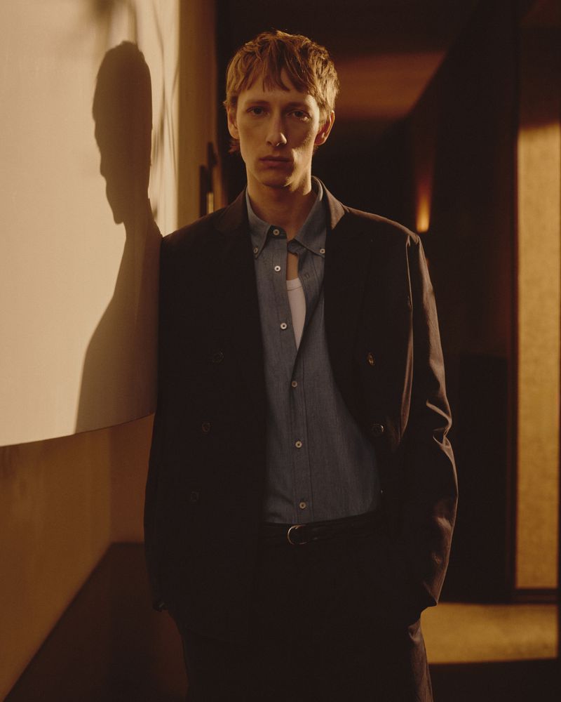 A stylish man in a suit stands indoors with a warm color cast, leaning against a wall, with a shadow cast on the wall behind him.