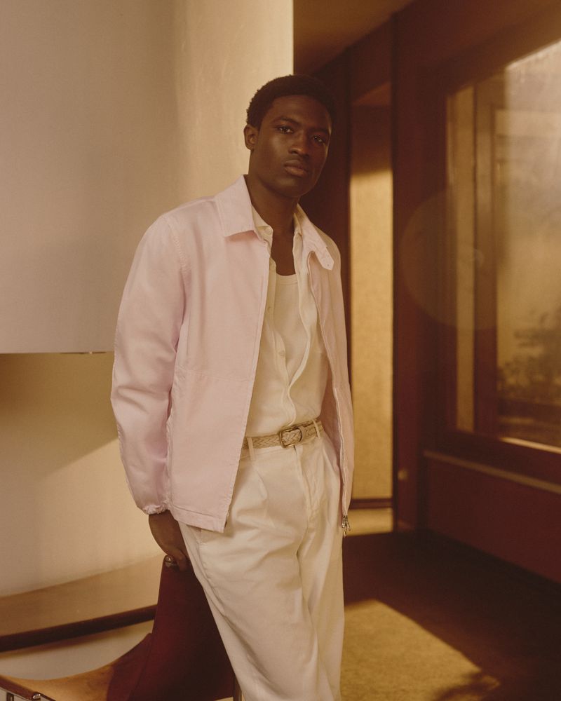 Indoor portrait of a Black man in a pink jacket and white pants, standing next to a chair, posing against a neutral wall.