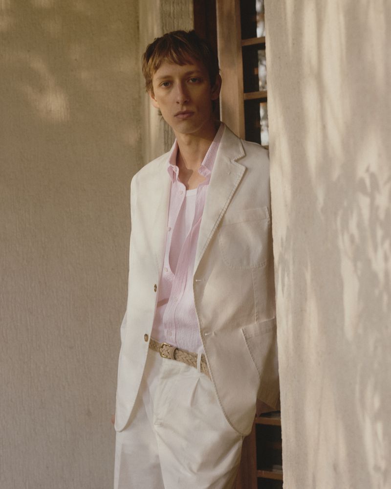 An adult male poses in a studio wearing a white suit and light pink button-down shirt while standing against a neutral wall.