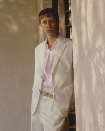 An adult male poses in a studio wearing a white suit and light pink button-down shirt while standing against a neutral wall.
