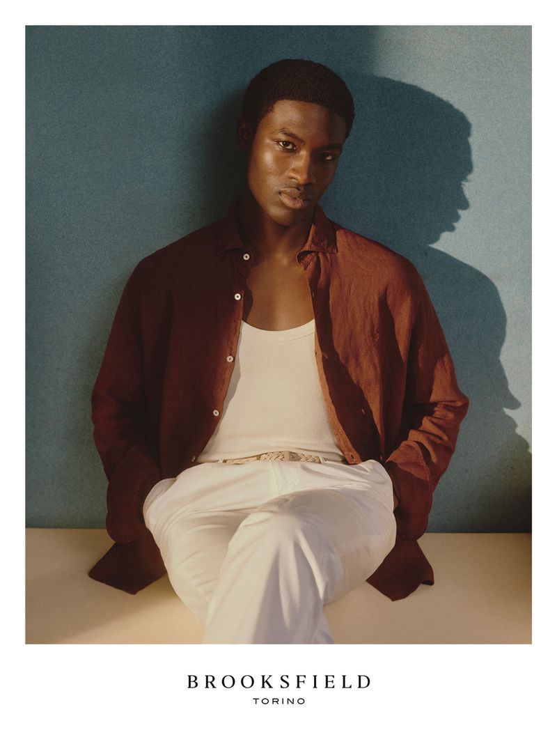 An adult male model with dark hair is wearing a brown button-down shirt, a white tank top, and white pants, posed against a textured blue wall.