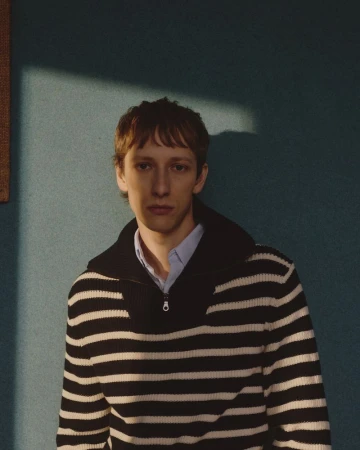 Close-up studio portrait of a young adult male with brown hair wearing a striped sweater and collared shirt against a teal background.
