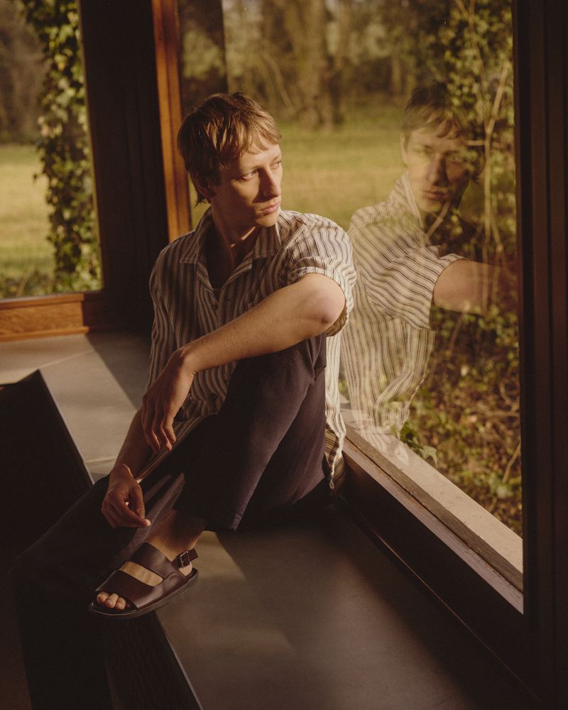 Man sits in front of a window with a reflection, wearing a striped shirt, sandals, and dark trousers, with a view of greenery outside.