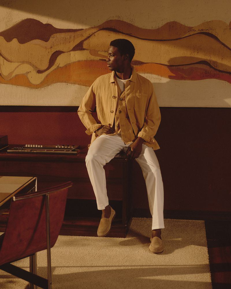 Black male model wearing a yellow jacket and white trousers seated indoors against a retro wall mural.