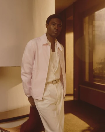 A Black male model posing indoors wearing a light pink jacket over white trousers for Brooksfield Spring Summer 2026 campaign