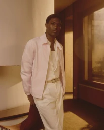 A Black male model posing indoors wearing a light pink jacket over white trousers for Brooksfield Spring Summer 2026 campaign