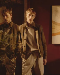 Male model wearing a brown jacket over a sweater and pale trousers standing indoors beside a mirror reflection