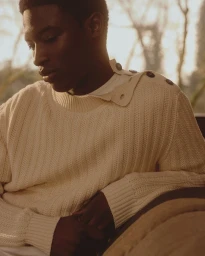 Close-up portrait of a Black man wearing a cream knit sweater with shoulder buttons outdoors in soft light