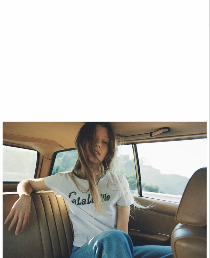 Young woman in a white t-shirt sitting in the back of a car with trees visible in the background through the car window.