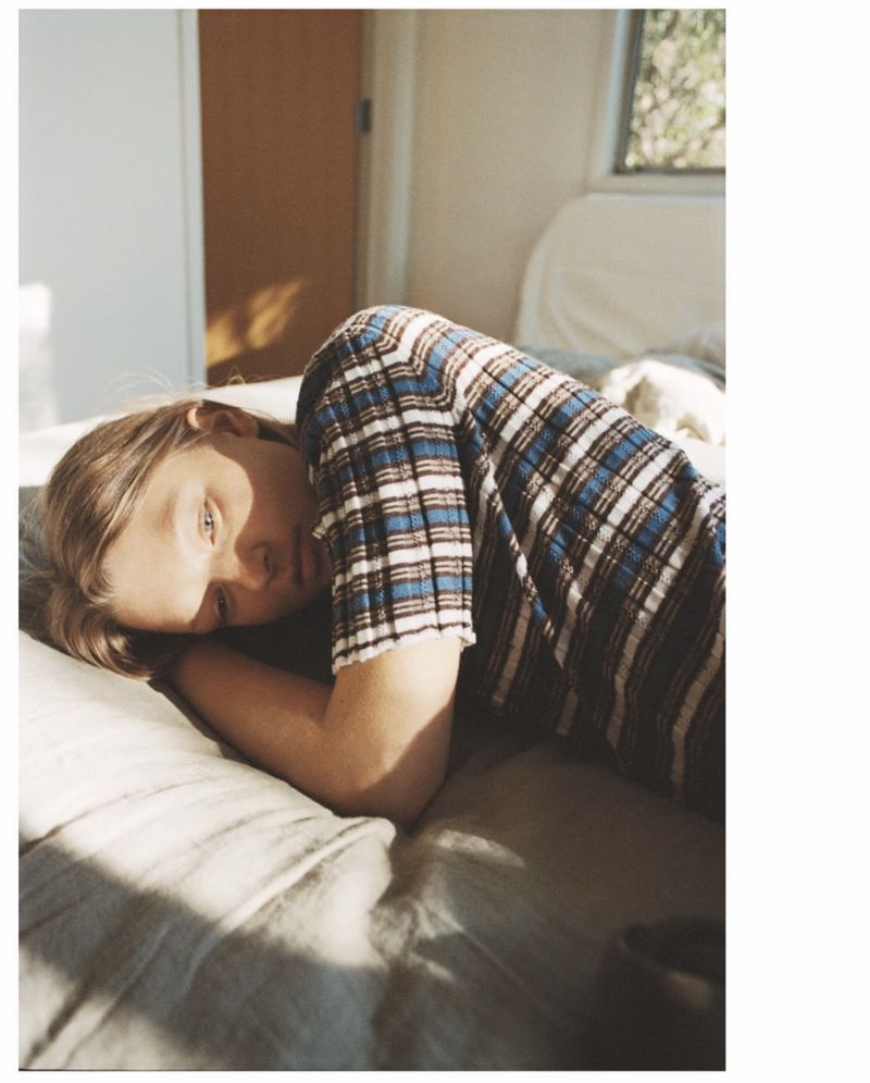 Young adult female lying on a bed in a bedroom with natural light, wearing a plaid shirt, in a comfortable and relaxed pose.