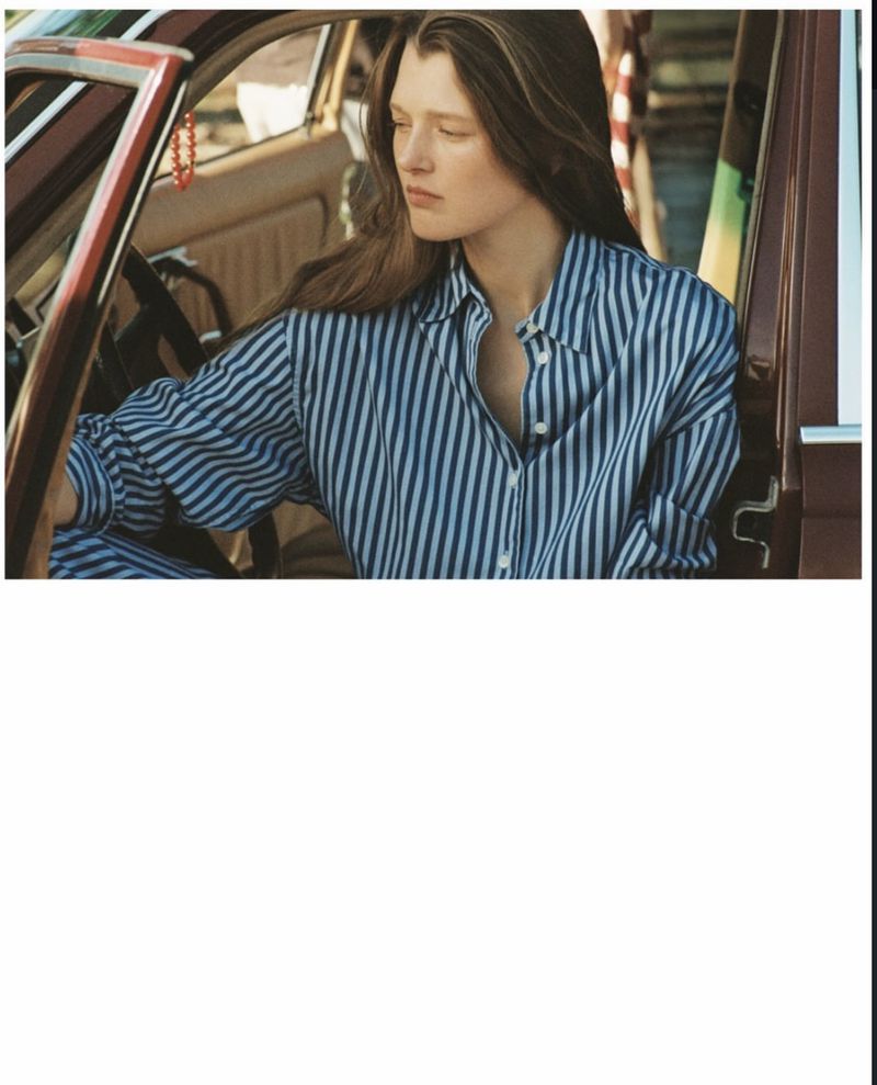 Young woman with long brown hair in blue and white striped shirt sitting in a vintage car with a reddish-brown interior.