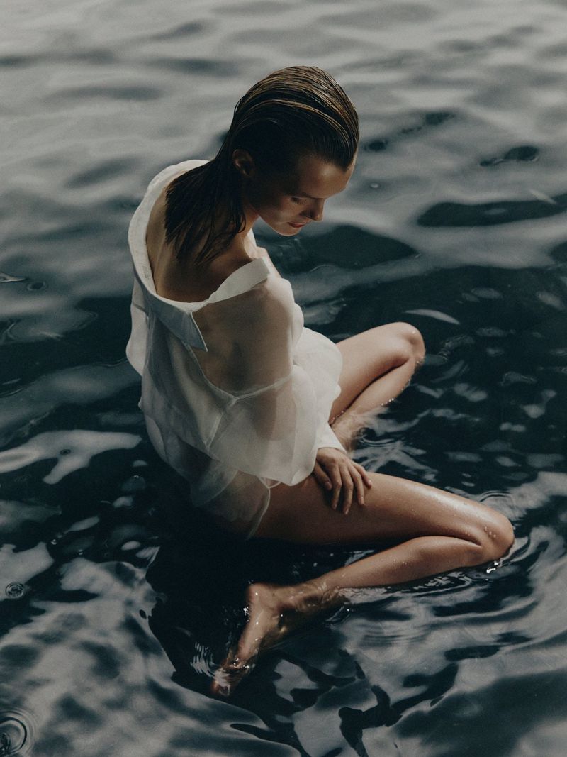 A woman sits in water, wearing a sheer white top and gazing downward, capturing a serene and reflective moment by the water's edge.