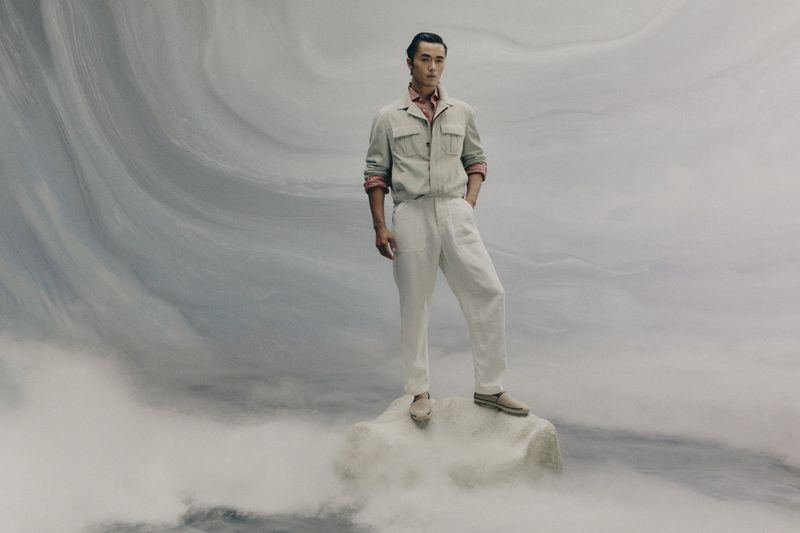 An Asian man stands confidently on a rock in a fashion shot. He is wearing a tan jacket, salmon shirt, and white pants against a hazy background.