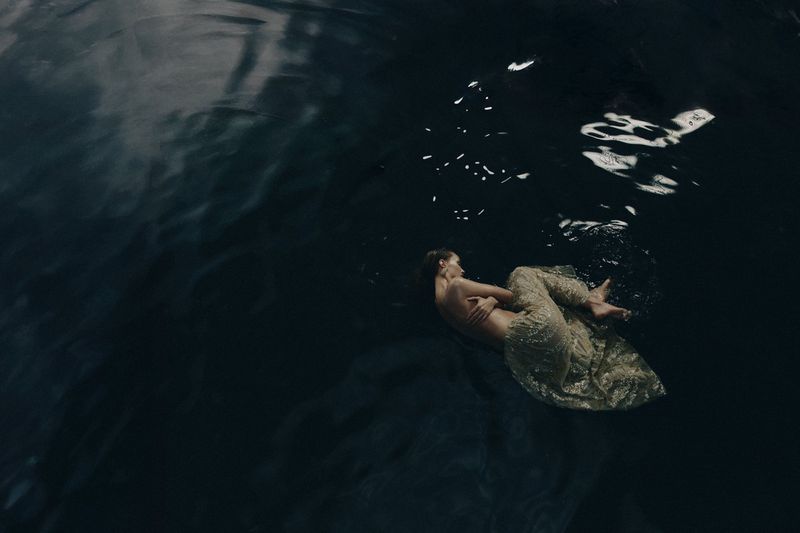 Overhead shot of a woman in a gold dress floating in dark, rippled water, creating a sense of calm and ethereal beauty.
