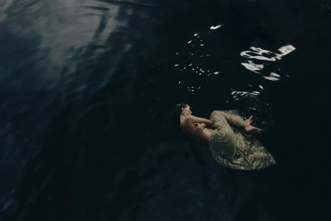 Overhead shot of a woman in a gold dress floating in dark, rippled water, creating a sense of calm and ethereal beauty.