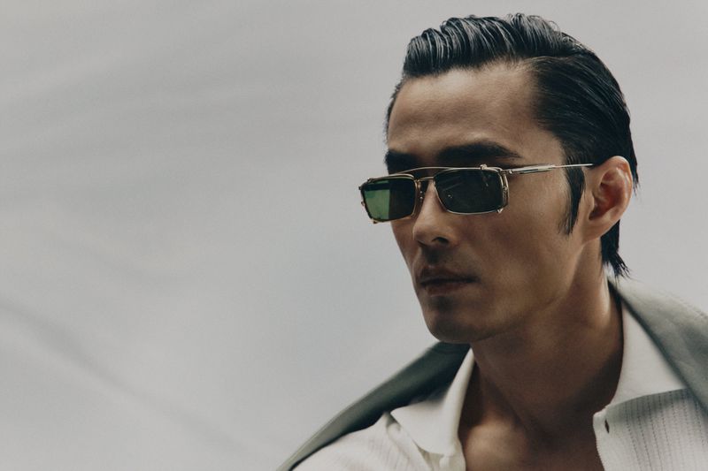 Portrait of an Asian man with slicked-back dark hair wearing green-lensed sunglasses, a white shirt, and a grey jacket.