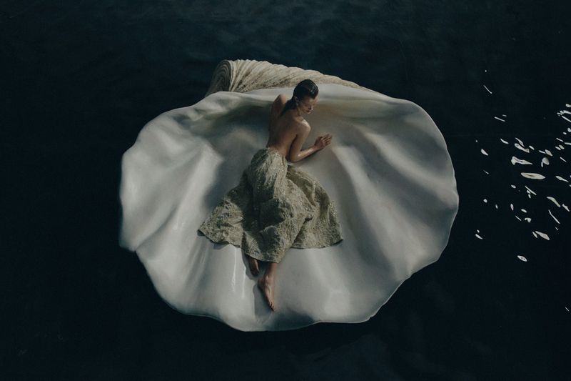 Aerial view of woman reclining in a giant seashell on water, wearing a textured dress, creating a Venus-like visual.