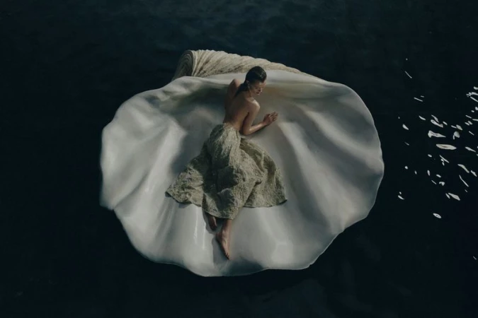 Aerial view of woman reclining in a giant seashell on water, wearing a textured dress, creating a Venus-like visual.