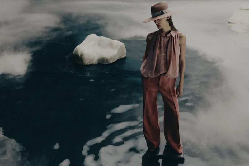 Fashionable adult female stands in water with a hat and stylish outfit, with fog in the background and a land formation.