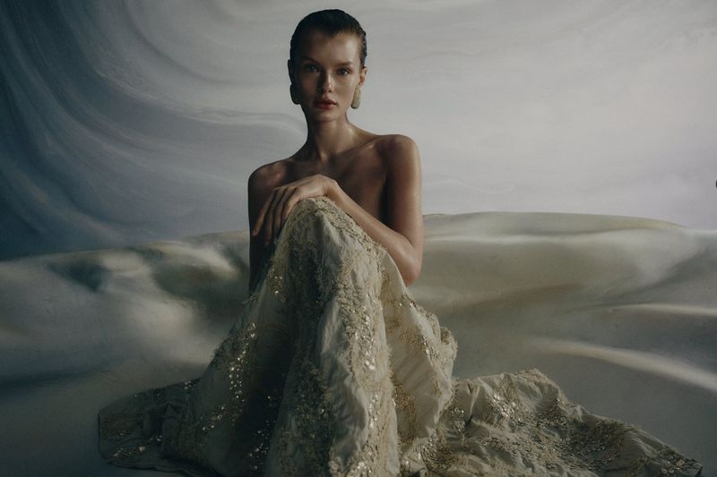 Model seated wearing an embellished cream skirt against a textured abstract backdrop for Brunello Cucinelli SS26.