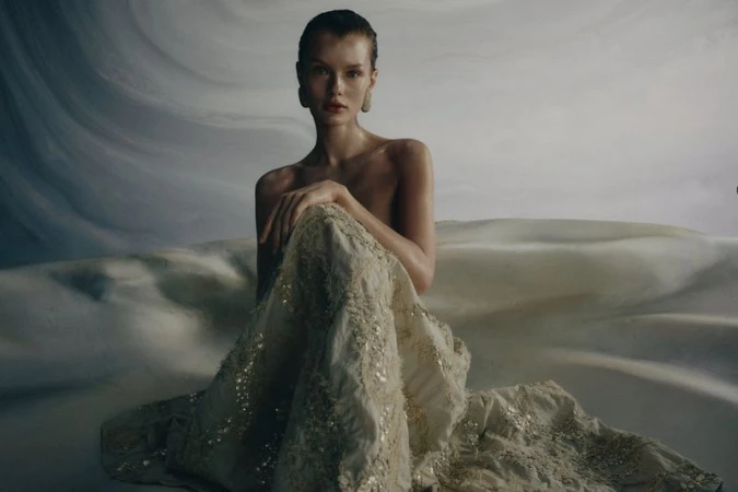 Model seated wearing an embellished cream skirt against a textured abstract backdrop for Brunello Cucinelli SS26.