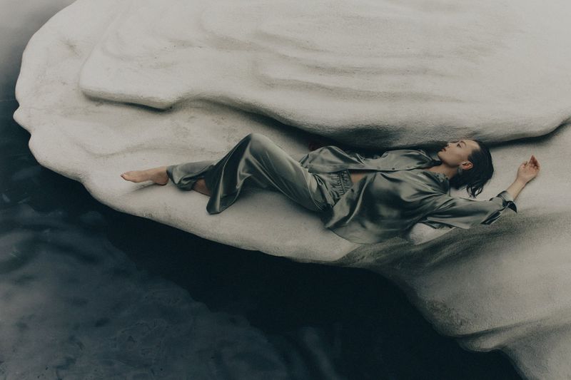 An adult female in a silk pajama set lies on a rock formation in water. The setting is natural with cool tones and soft lighting.