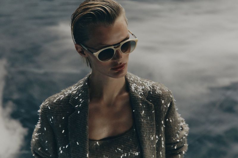 Adult female model with short blond hair poses in a jacket and top with glitter accents against a blurred grey cloud background indoors.