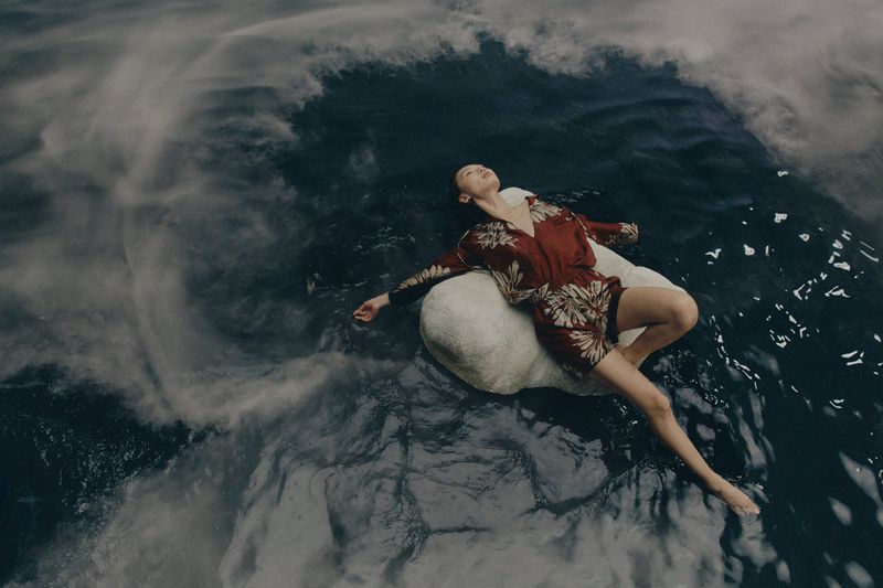 An adult woman in a floral-patterned playsuit is reclining on a white pillow or soft boulder in a body of dark rippled water.