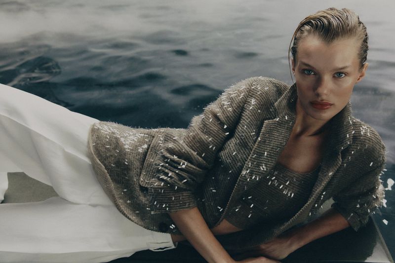 Adult female fashion model poses in a beige tweed-like blazer and top while lying on a white surface with water in the background.