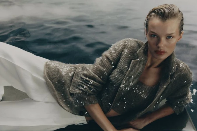 Adult female fashion model poses in a beige tweed-like blazer and top while lying on a white surface with water in the background.