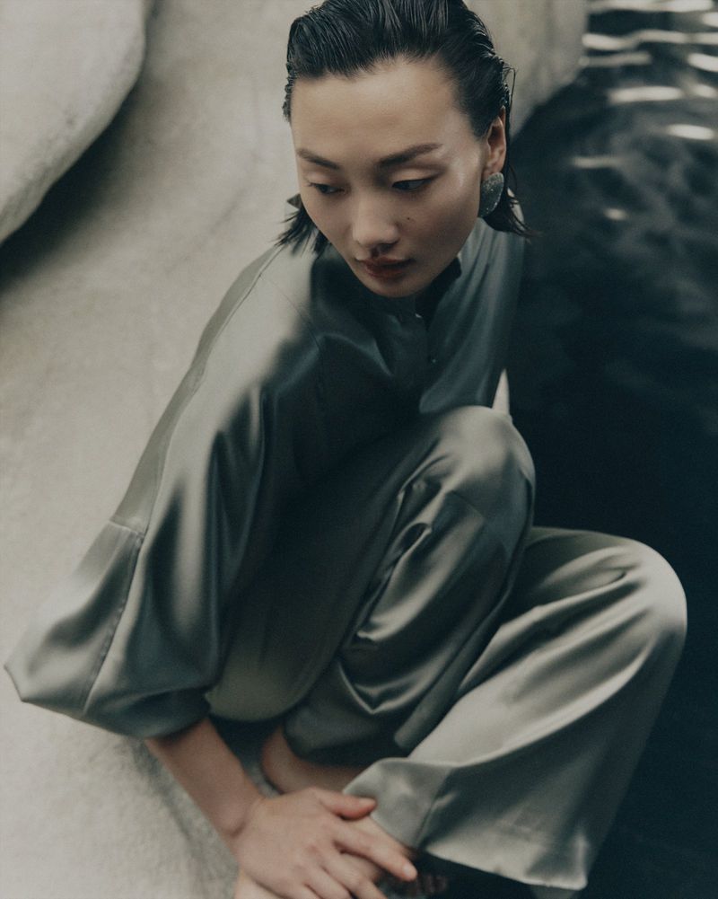 Adult woman in a crouched pose wearing a silky green outfit by a light-colored rock formation and dark water. She has dark hair and bare feet.