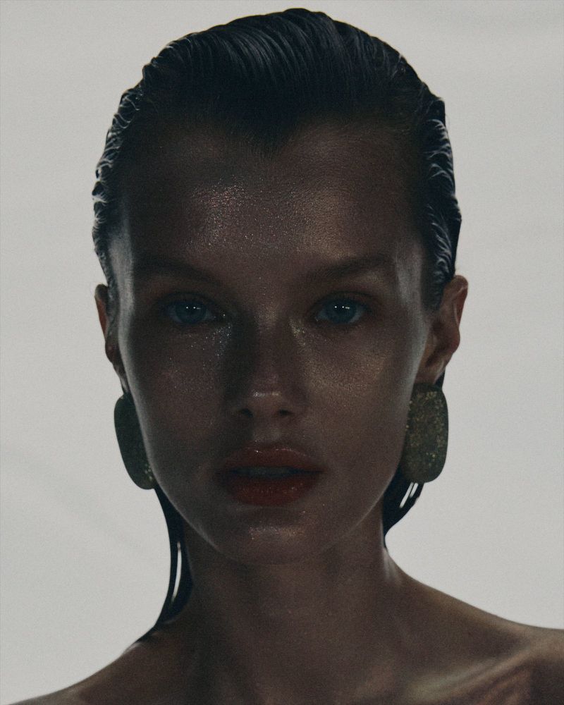 Close-up portrait of an adult female with slicked-back dark hair, large oval earrings, and red lipstick against a bright white backdrop.