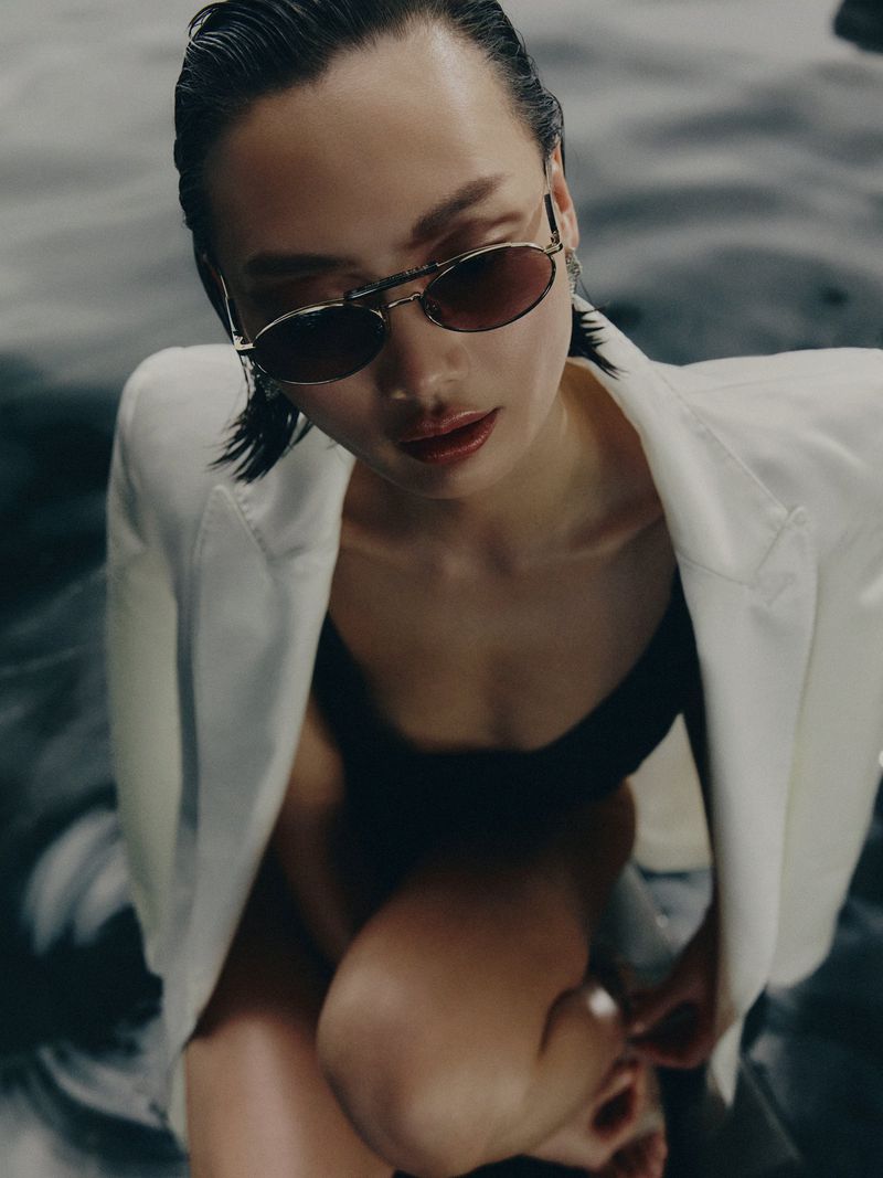 Adult female model with slicked-back hair and sunglasses in water, wearing a white blazer and black top, posing for fashion photography.