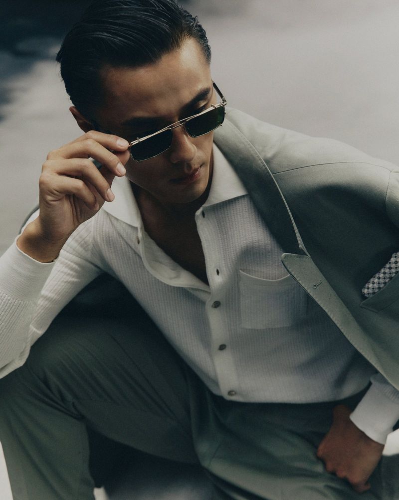 Attractive Asian man poses in sunglasses and a suit, sitting indoors with soft studio light, showcasing modern fashion and sophisticated style.