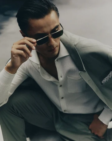 Attractive Asian man poses in sunglasses and a suit, sitting indoors with soft studio light, showcasing modern fashion and sophisticated style.